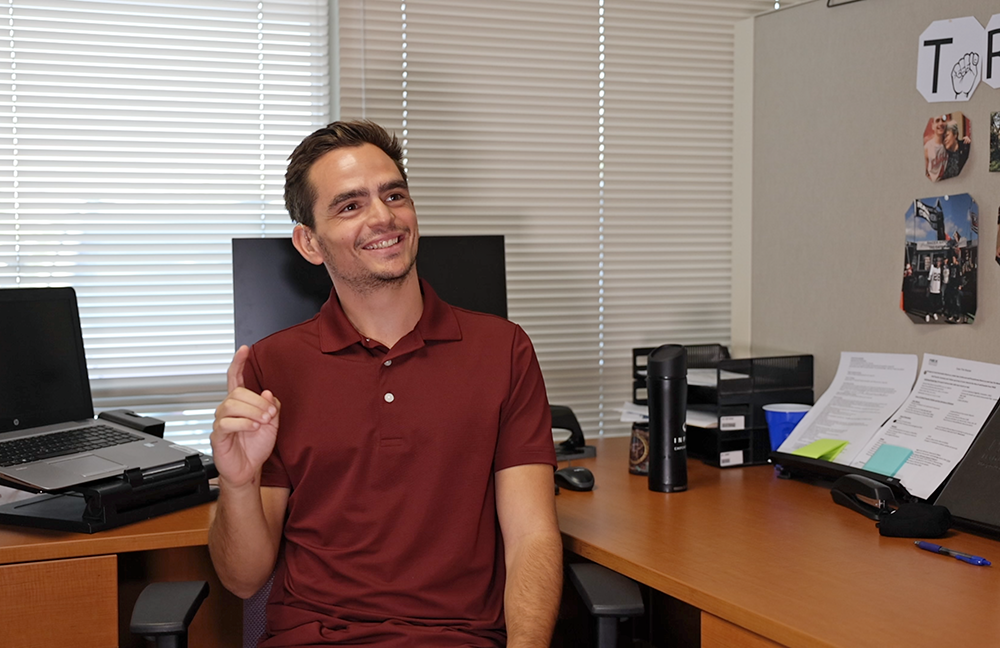 image of man in red shirt at desk smiling and doing sign language