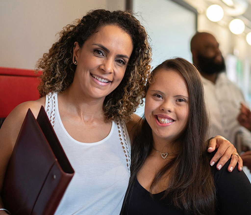 image of woman in white tank top holding portfolio folder smiling with her arm around a woman with down syndrome smiling