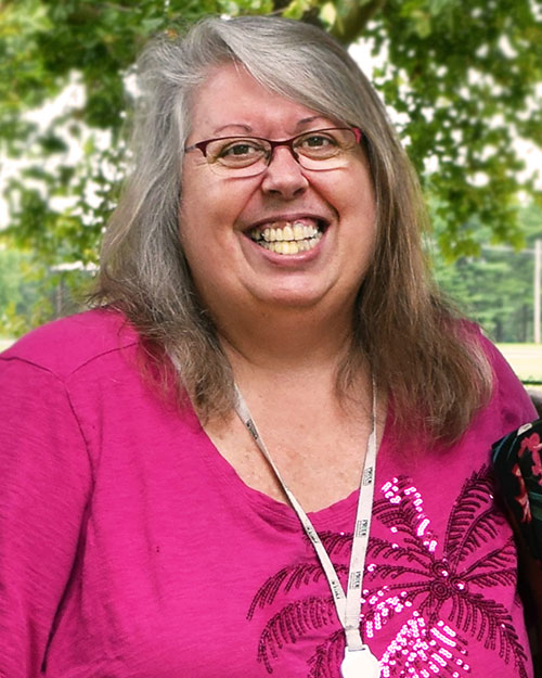 woman with medium length gray hair with glasses wearing a pink shirt smiling