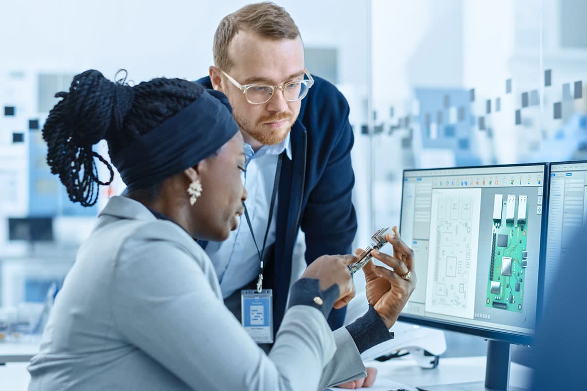 Two engineers, one standing, one seated at a desk, review a board design