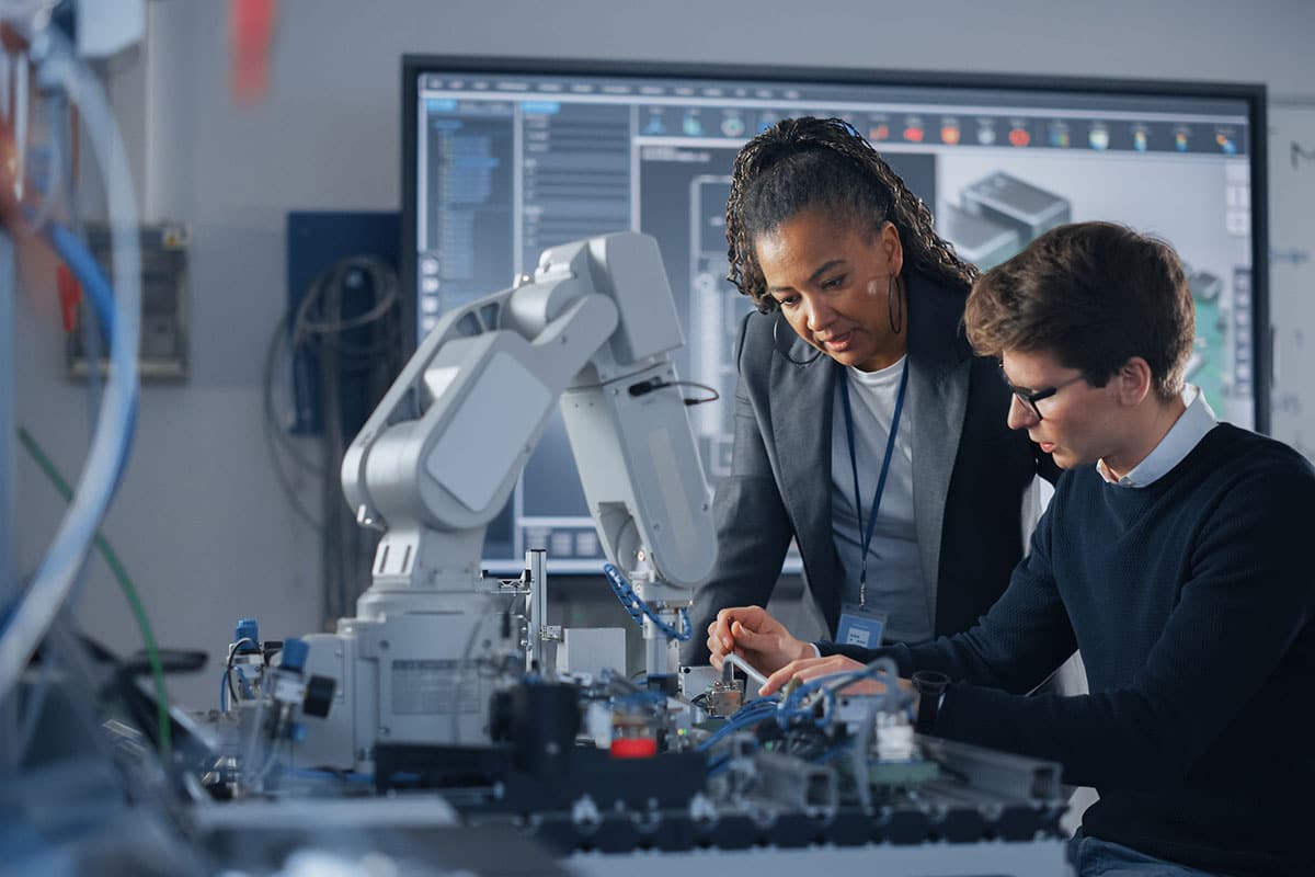 A pair of engineers inspecting a prototype, with a large schematic on a display screen behind them