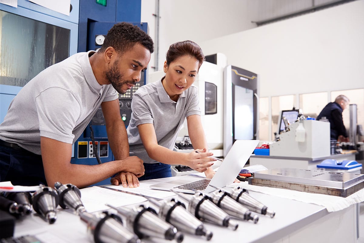 A man and a woman, standing at a desk in a manufacturing facility, looking at a laptop computer screen.