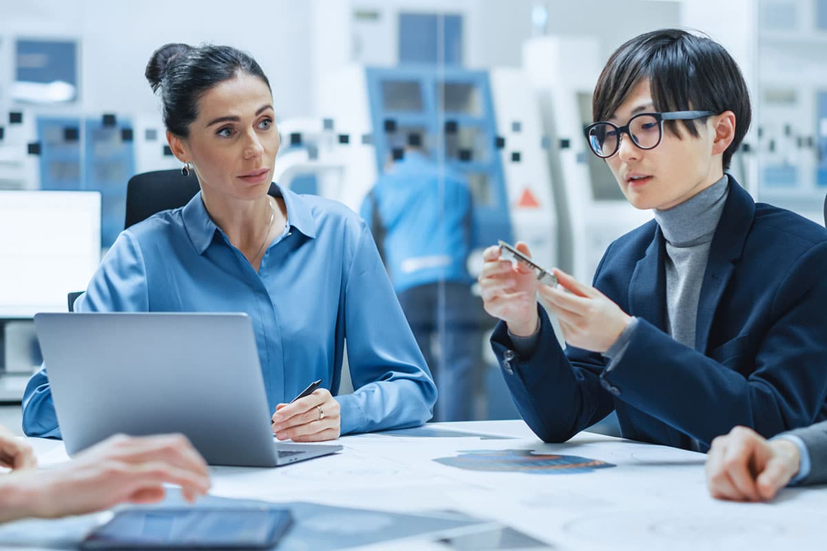 A woman and a man in glasses seated at a table in a manufacturing facility, examining one of the components of an electronics device.