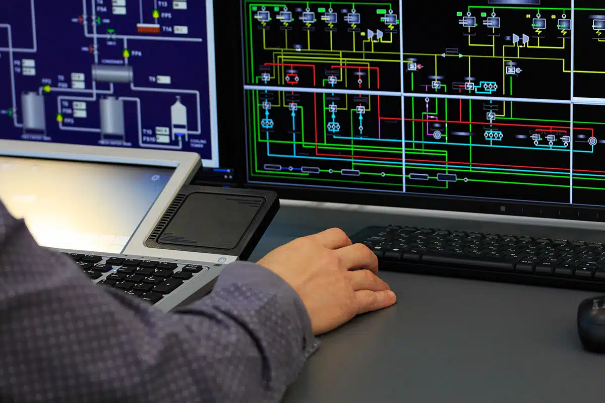 A close-up of a man’s hand using a computer mouse, with a building schematic showing on a computer screen