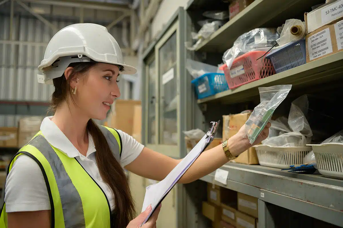 A young woman in hardhat and safety vest, holding a clipboard and standing in front of shelves holding electronics components