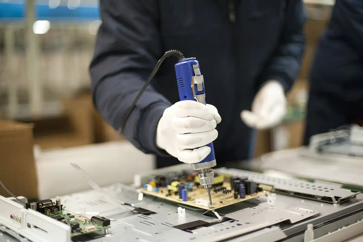 A closeup of a technician in an electronics factory, wearing latex gloves, using an electric screwdriver to attach a PCBA to a product’s shell