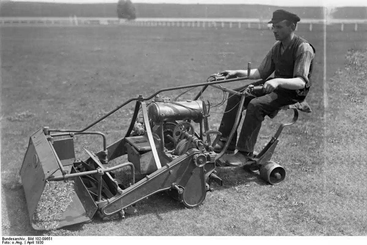 A black-and-white photo of a man, wearing a vest and cap, sitting on a large, mechanical mower