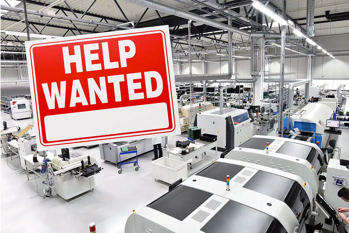 A Help Wanted sign affixed to a photo of the factory floor of an electronics manufacturing company