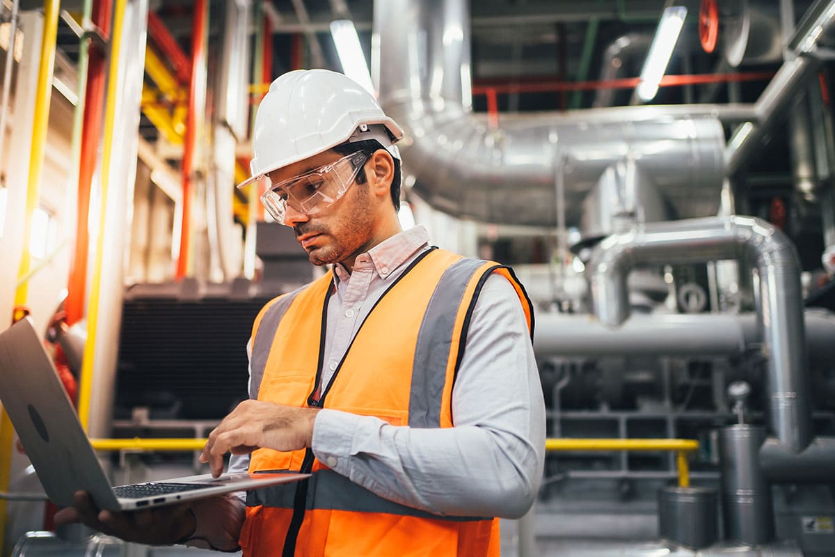 A technician in hardhat, goggles, and safety vest, standing in front of an industrial HVAC configuration, looking at a laptop