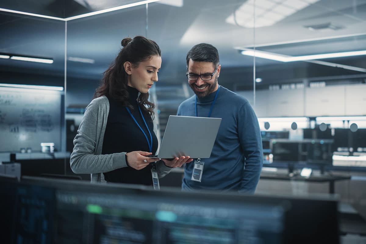 Two engineers, a woman and a man, standing in a glassed-in conference room, looking at a laptop computer the woman is holding