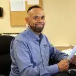 Image of Hispanic man in PRIDE Industries collared shirt sitting at desk smiling