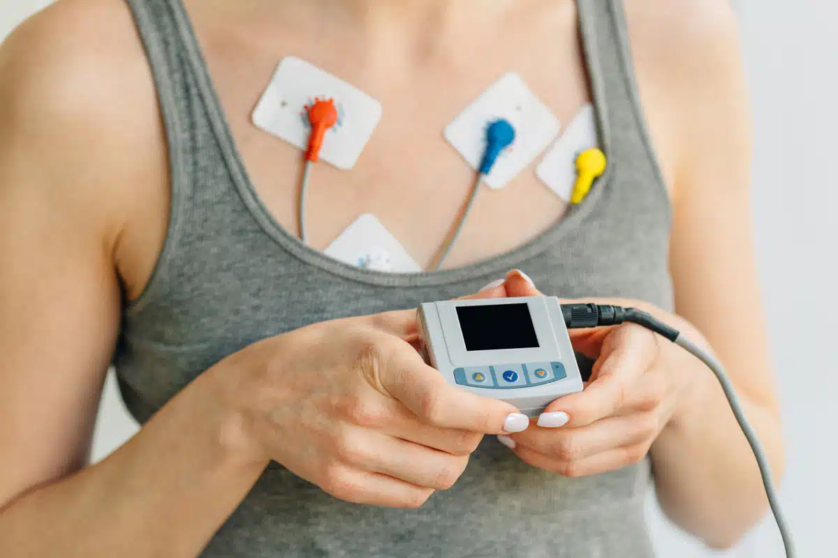 A closeup of a woman in a tank top, with diagnostic leads on her chest that are attached to a monitoring device she’s holding