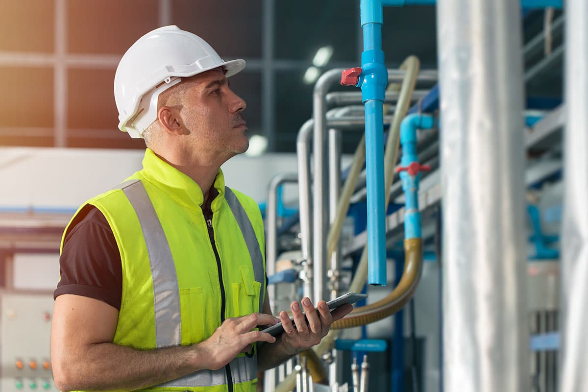 A technician wearing a hard hat and safety vest, holding a tablet, examines facility equipment