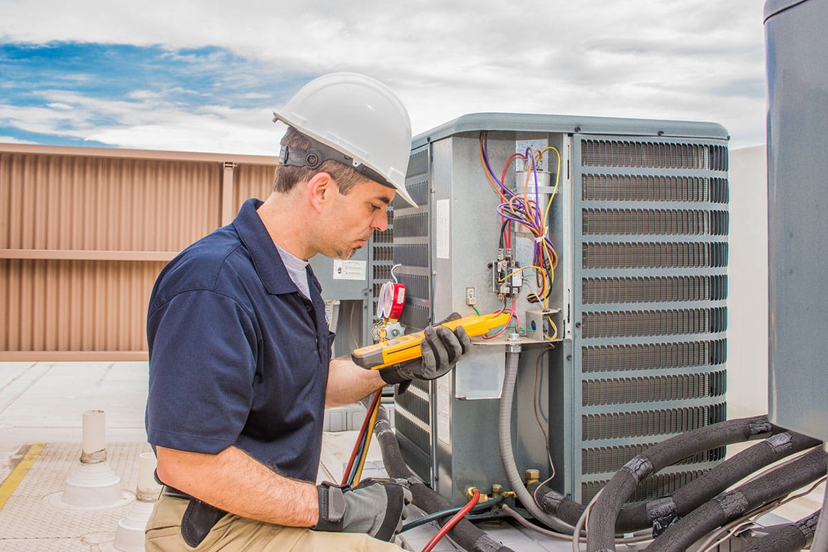 A man wearing a hardhat uses a handheld device to check an outdoor component of an HVAC system.