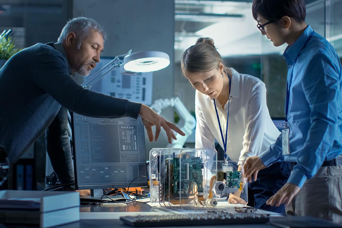 An electronics engineer talking with two customers as they stand at a worktable looking at a prototype electronic device