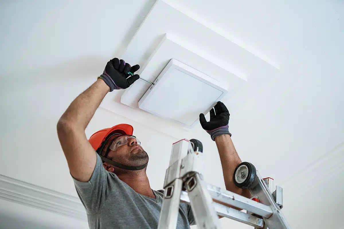 A man standing on a ladder, wearing a hardhat, goggles, and gloves, uses a screwdriver to remove the cover of a light fixture.