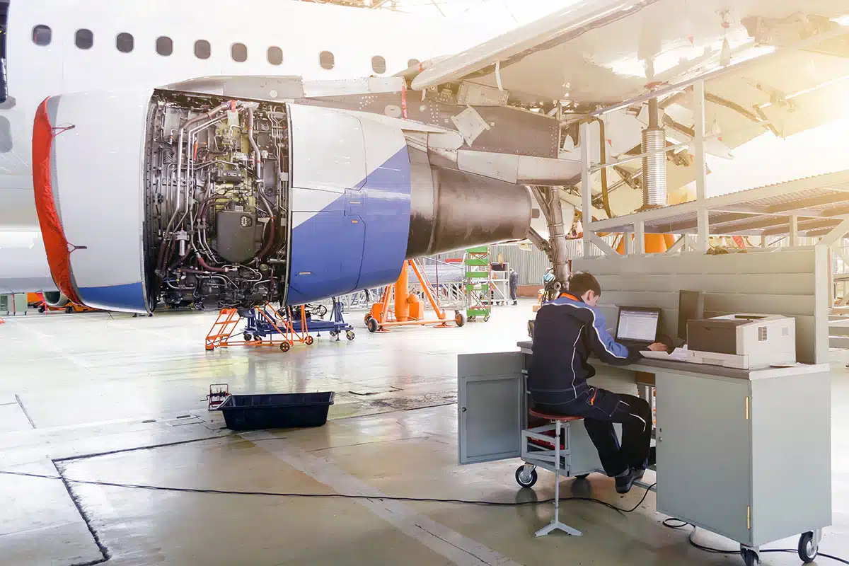 A technician at a workstation in an aircraft hangar, next to an airplane with the cowling removed from one engine.