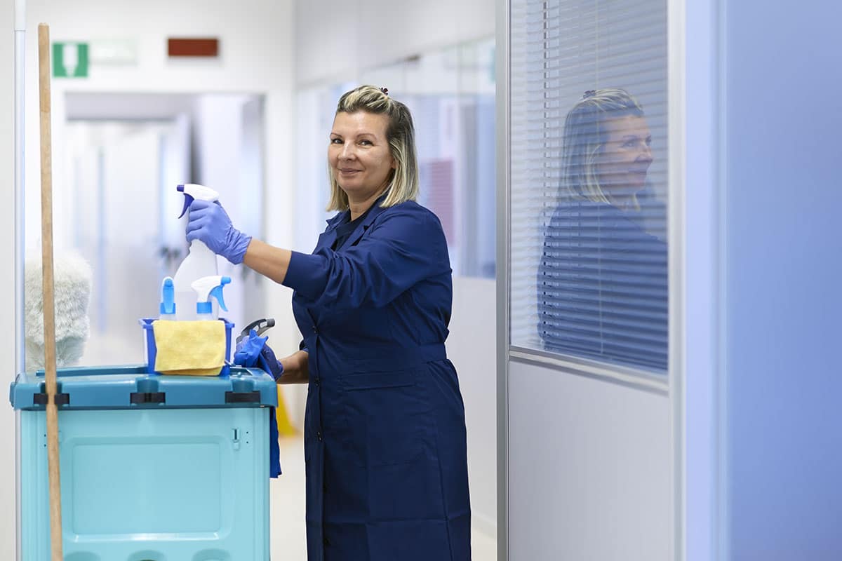 A woman wearing gloves and a hospital custodial uniform, standing next to a cleaning cart, holding a spray bottle.