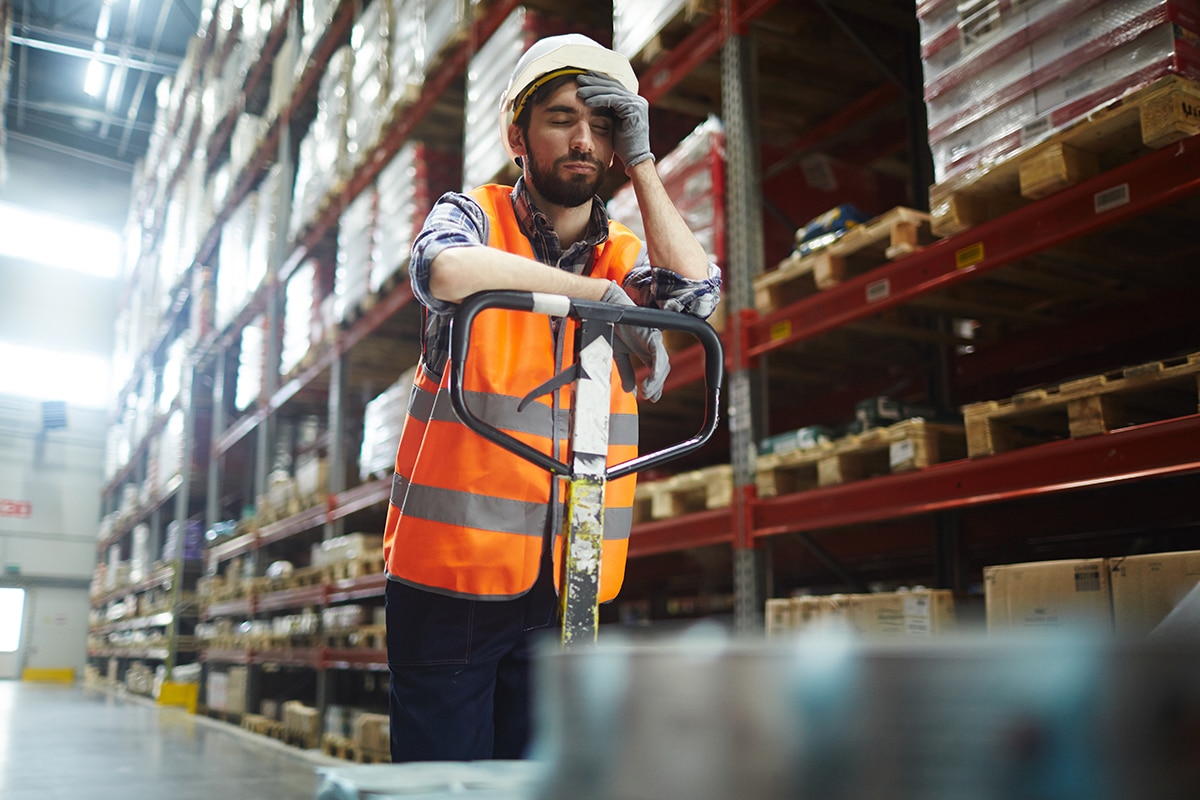 : A worker in a hardhat and orange vest, looking bored, leans against a piece of equipment