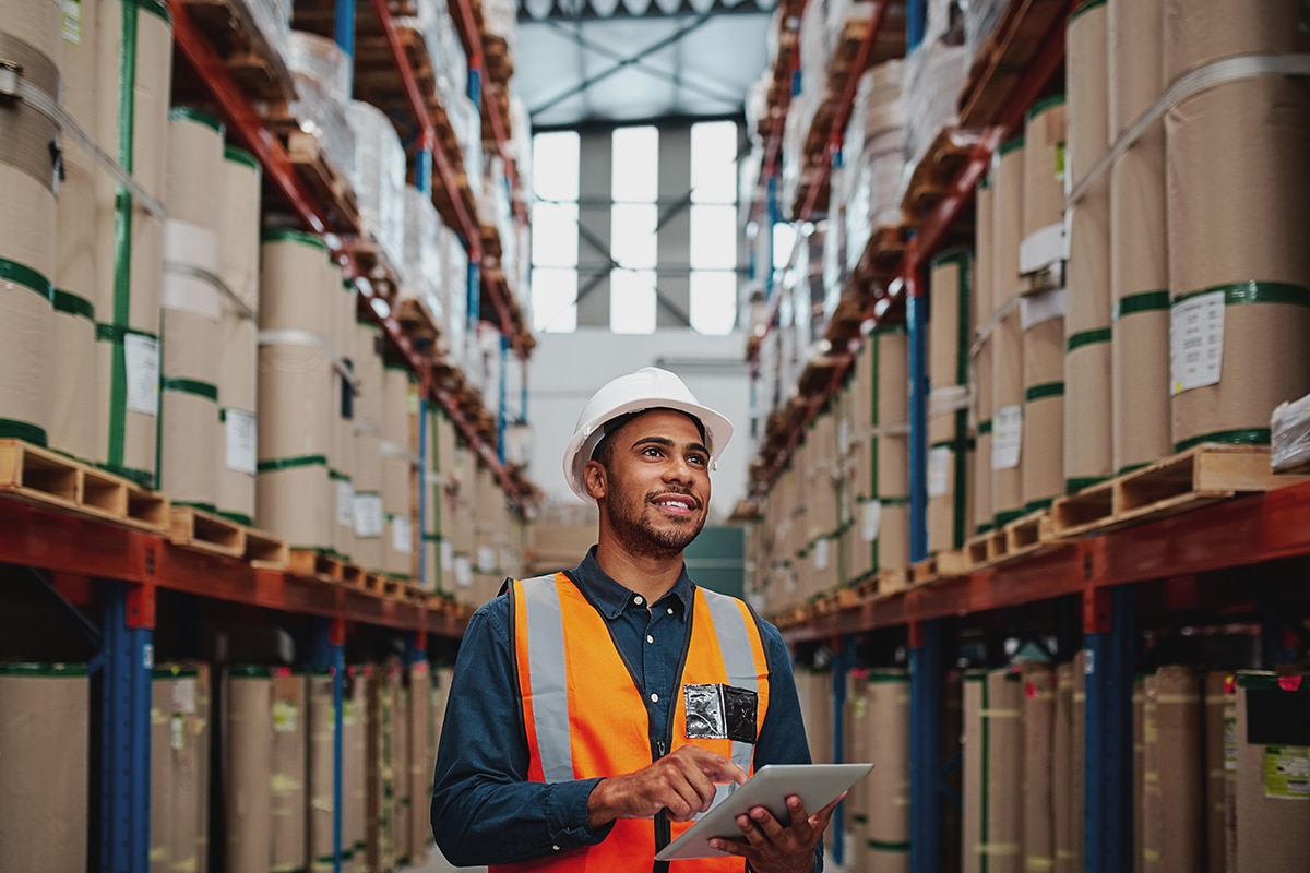 A warehouse worker stands in the middle of a warehouse, using a handheld device and looking up at stacks of products.