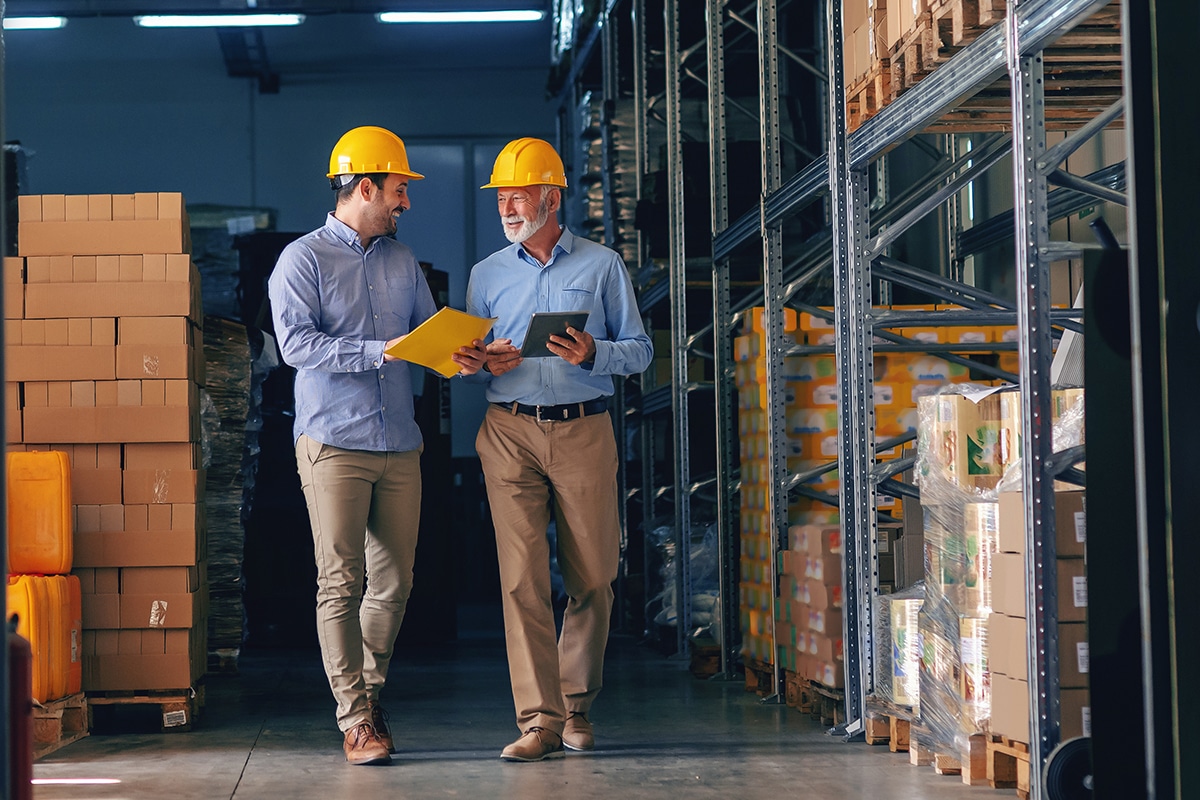 Two men, wearing dress shirts and hardhats, look at paperwork as they walk together through a warehouse.