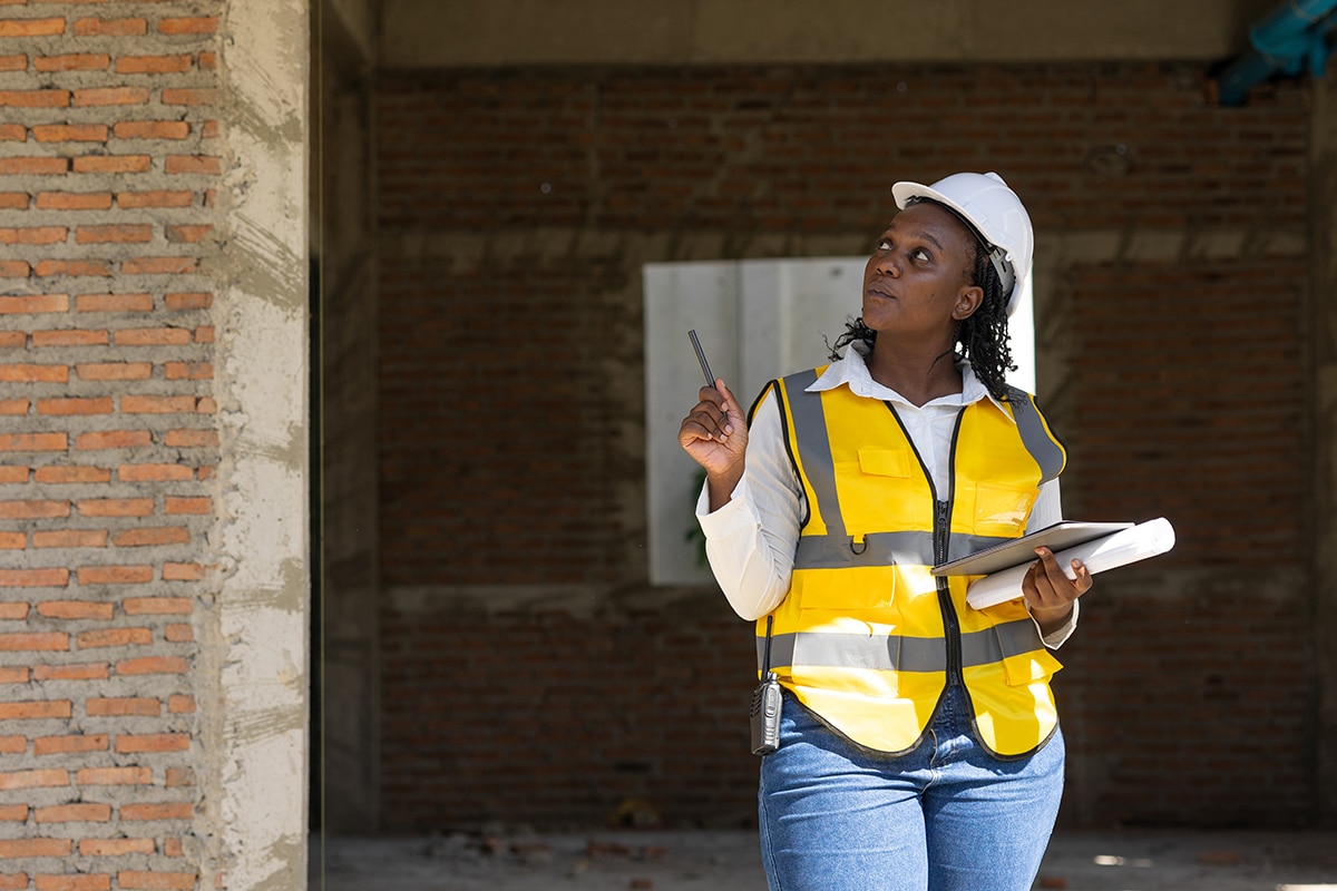 A woman in a safety vest and hardhat examines the brick exterior of a building.