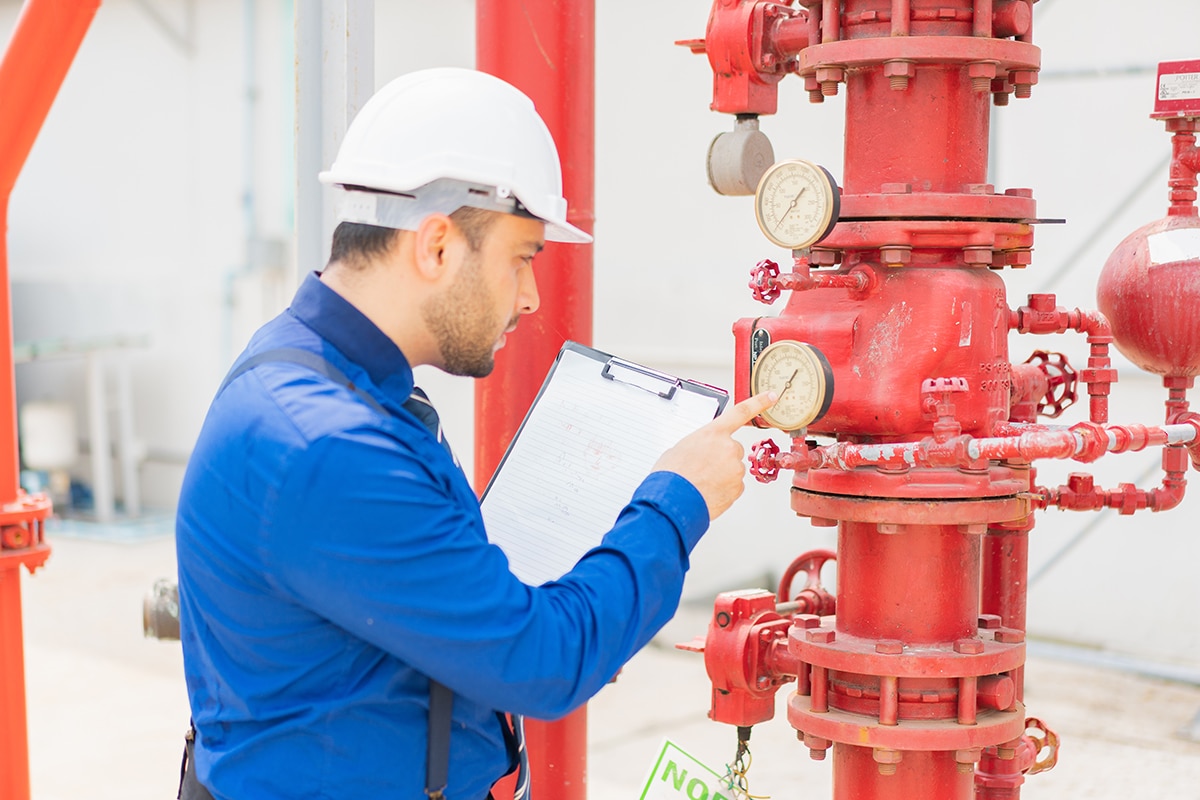 A man in a dress shirt and hardhat, holding a clipboard, gets a reading from a gauge attached to a large red pipe.