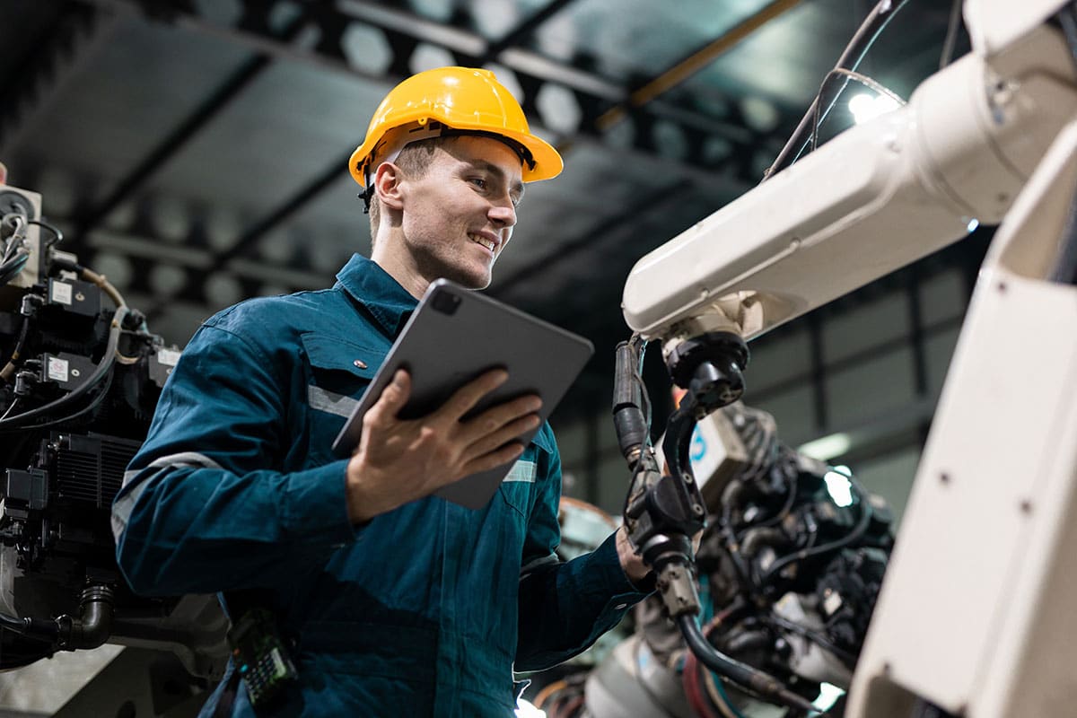 A man in a hardhat and work overalls, holding a computer tablet, adjusts a setting on an industrial robot.