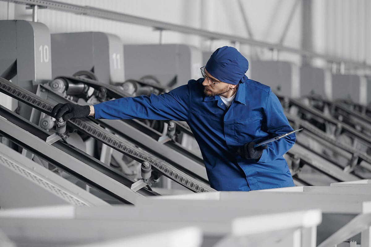 A man in a blue work smock and hair cover inspects a piece of equipment in a high-tech factory.