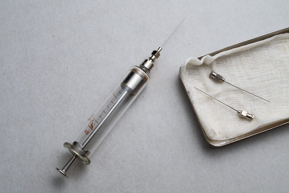 An old-fashioned glass-and-metal syringe lies next to a tray holding gauze and steel needles.